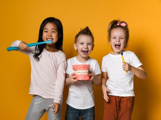 Two smiling kids holding large fake toothbrushes with another kid holding model of the mouth